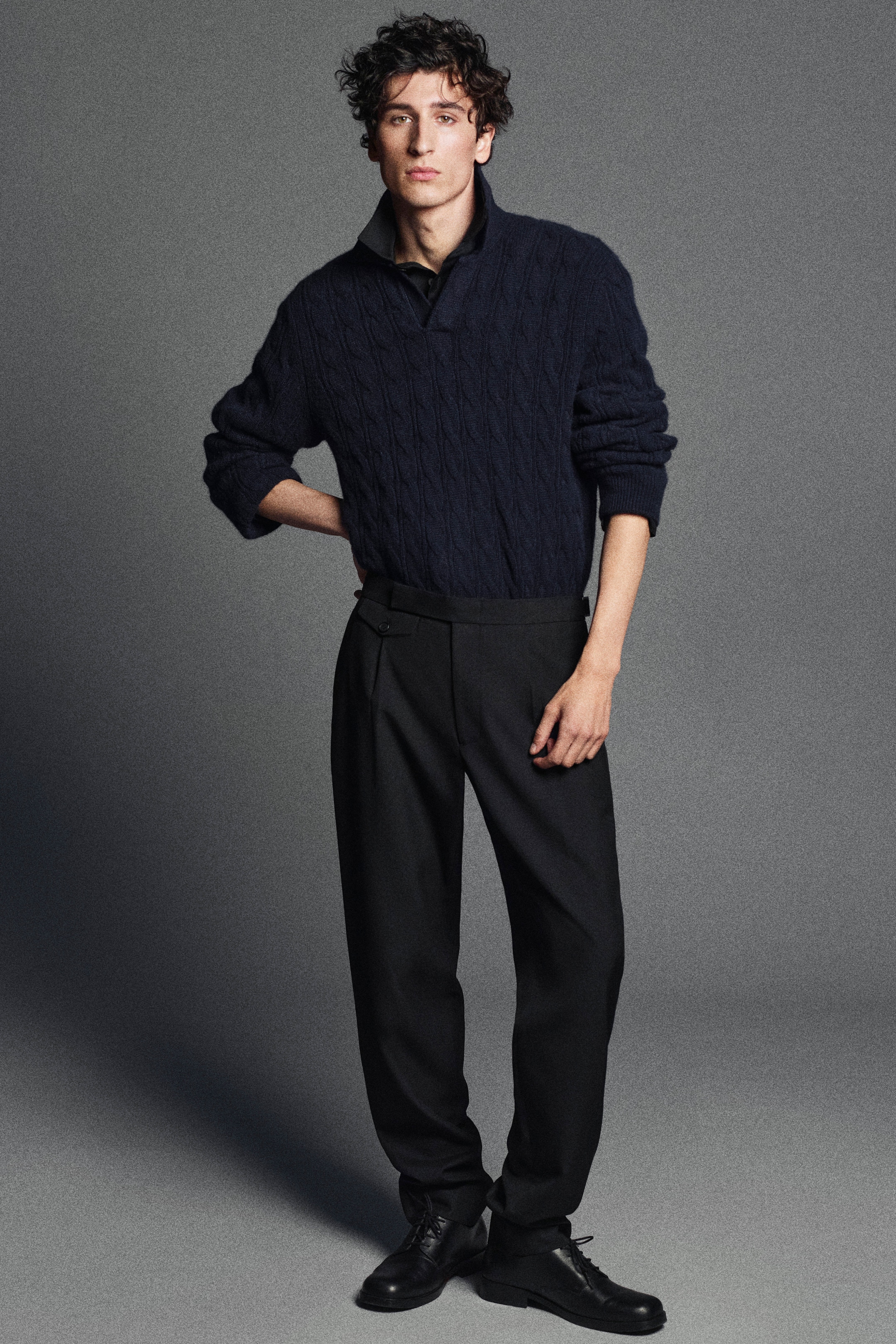 A person with curly hair wearing a dark cable-knit sweater, dark trousers, and black shoes stands against a gray background.
