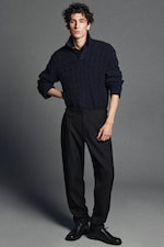 Placeholder for: A person with curly hair wearing a dark cable-knit sweater, dark trousers, and black shoes stands against a gray background.