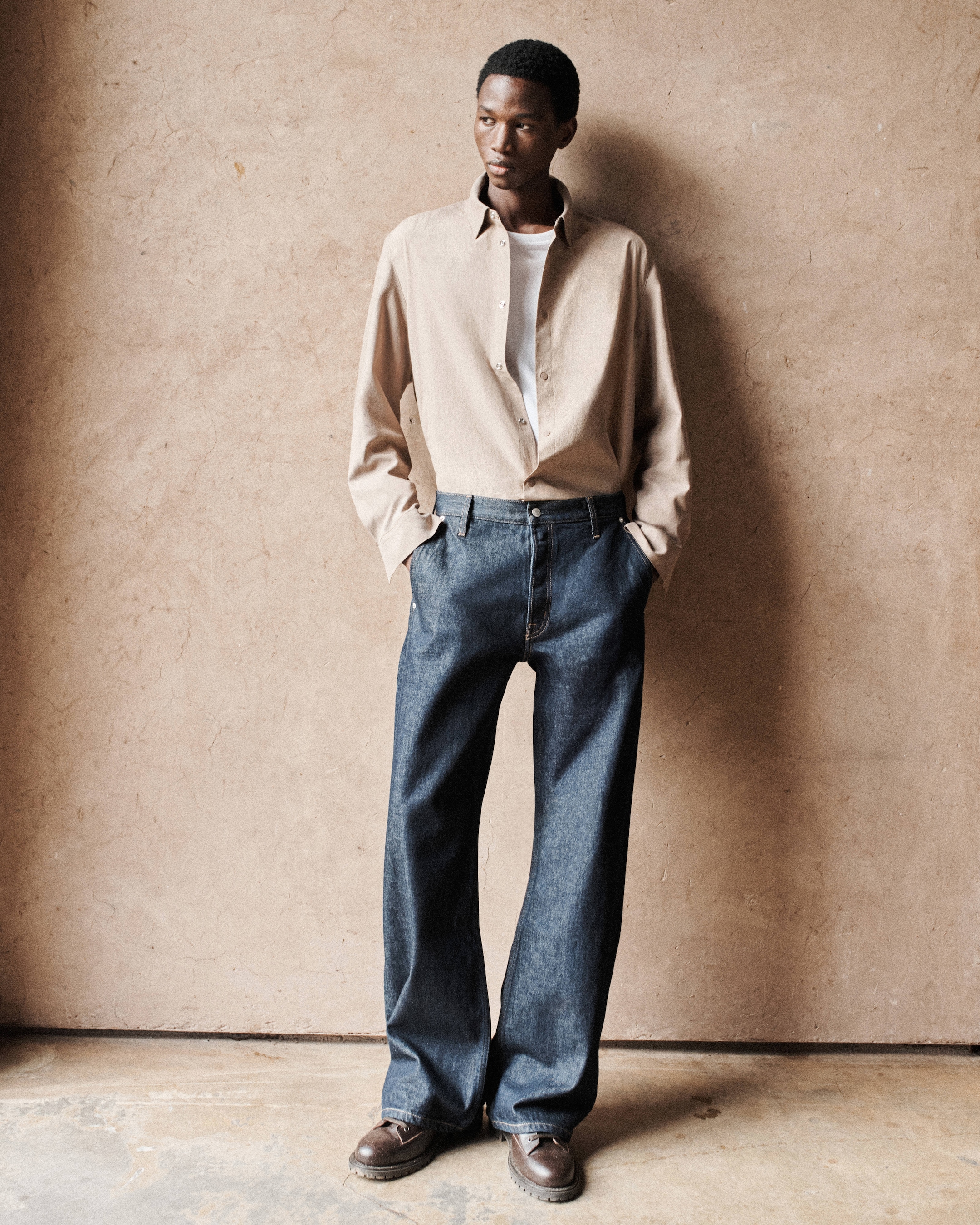 Person standing against a textured wall, wearing a beige shirt, white undershirt, blue jeans, and brown shoes, hands in pockets.