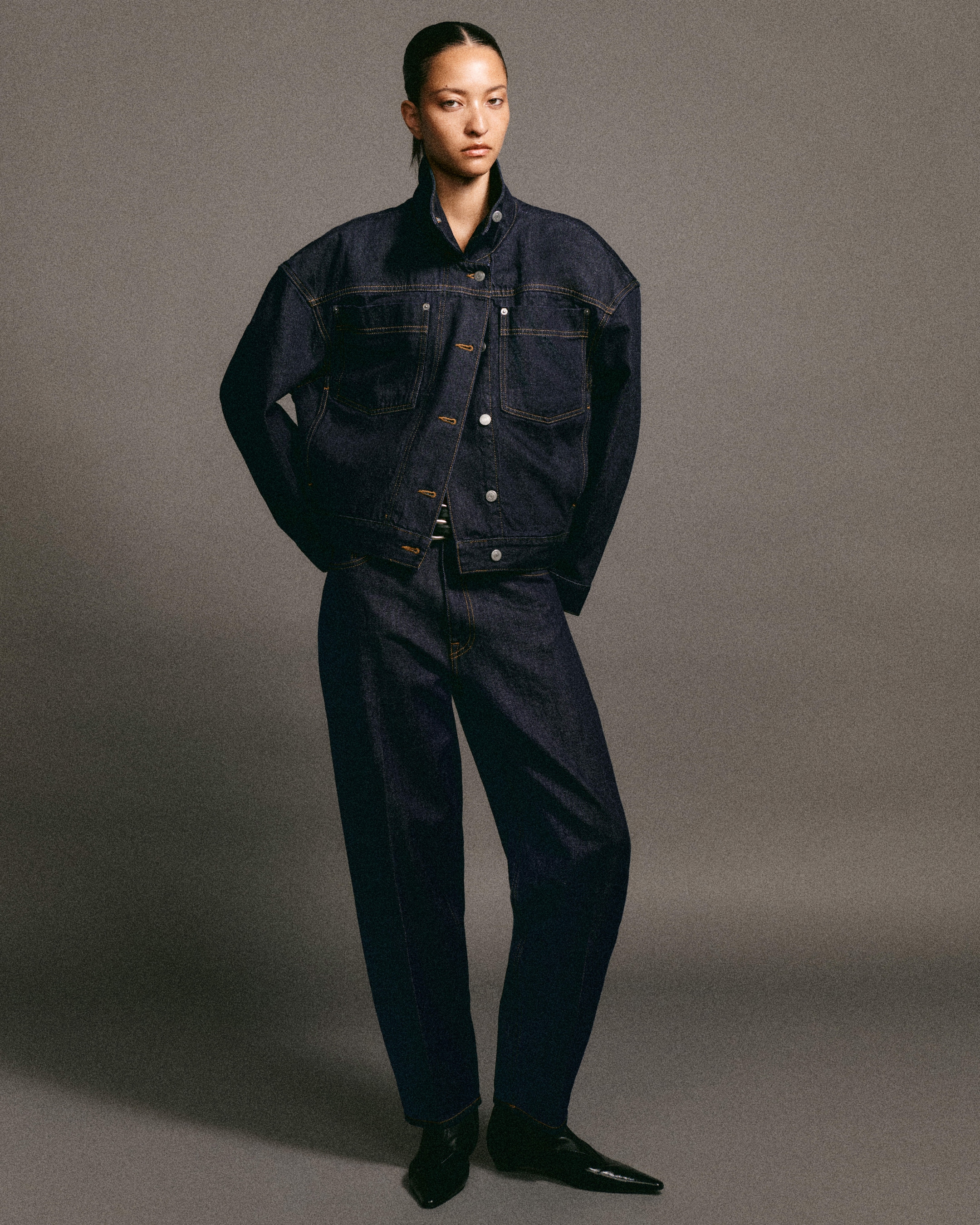 A person is wearing a dark blue denim jacket and jeans, posing confidently against a neutral gray background.