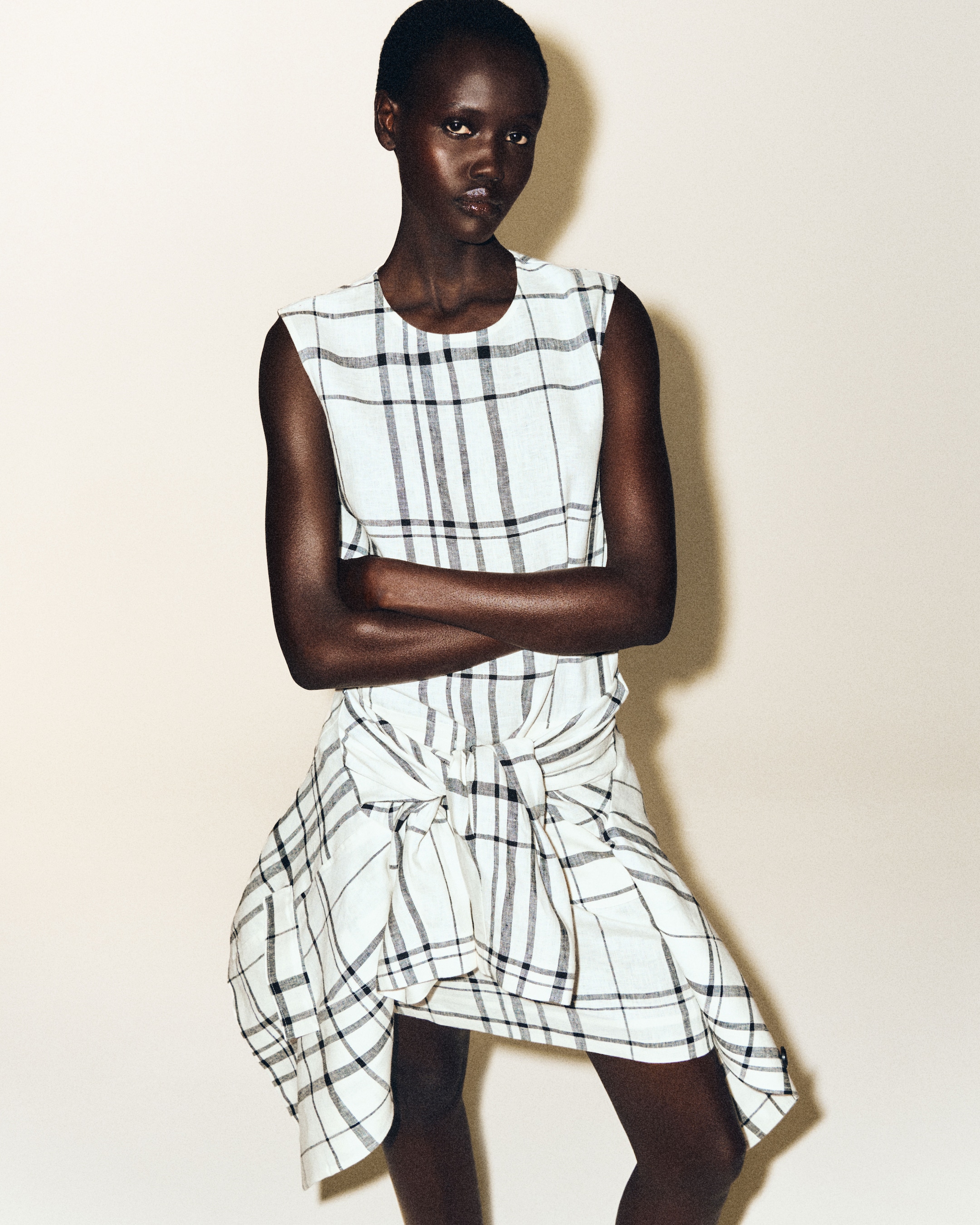 A person wearing a sleeveless plaid dress with arms crossed, standing against a neutral background.