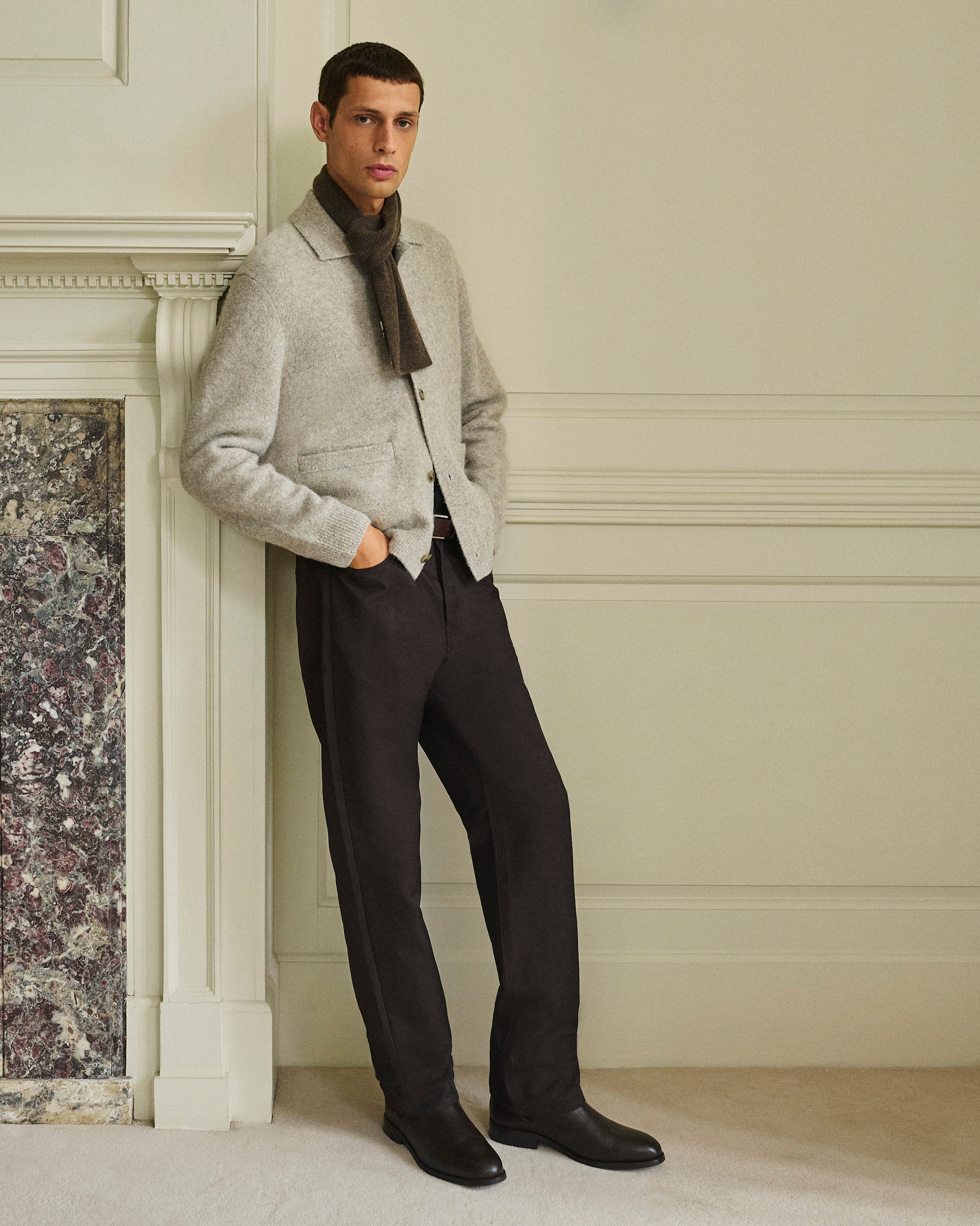 A man wearing a light sweater, scarf, dark trousers, and boots stands against a wall with hands in pockets, next to a decorative fireplace.
