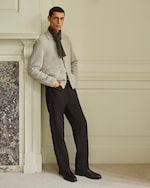 Placeholder for: A man wearing a light sweater, scarf, dark trousers, and boots stands against a wall with hands in pockets, next to a decorative fireplace.