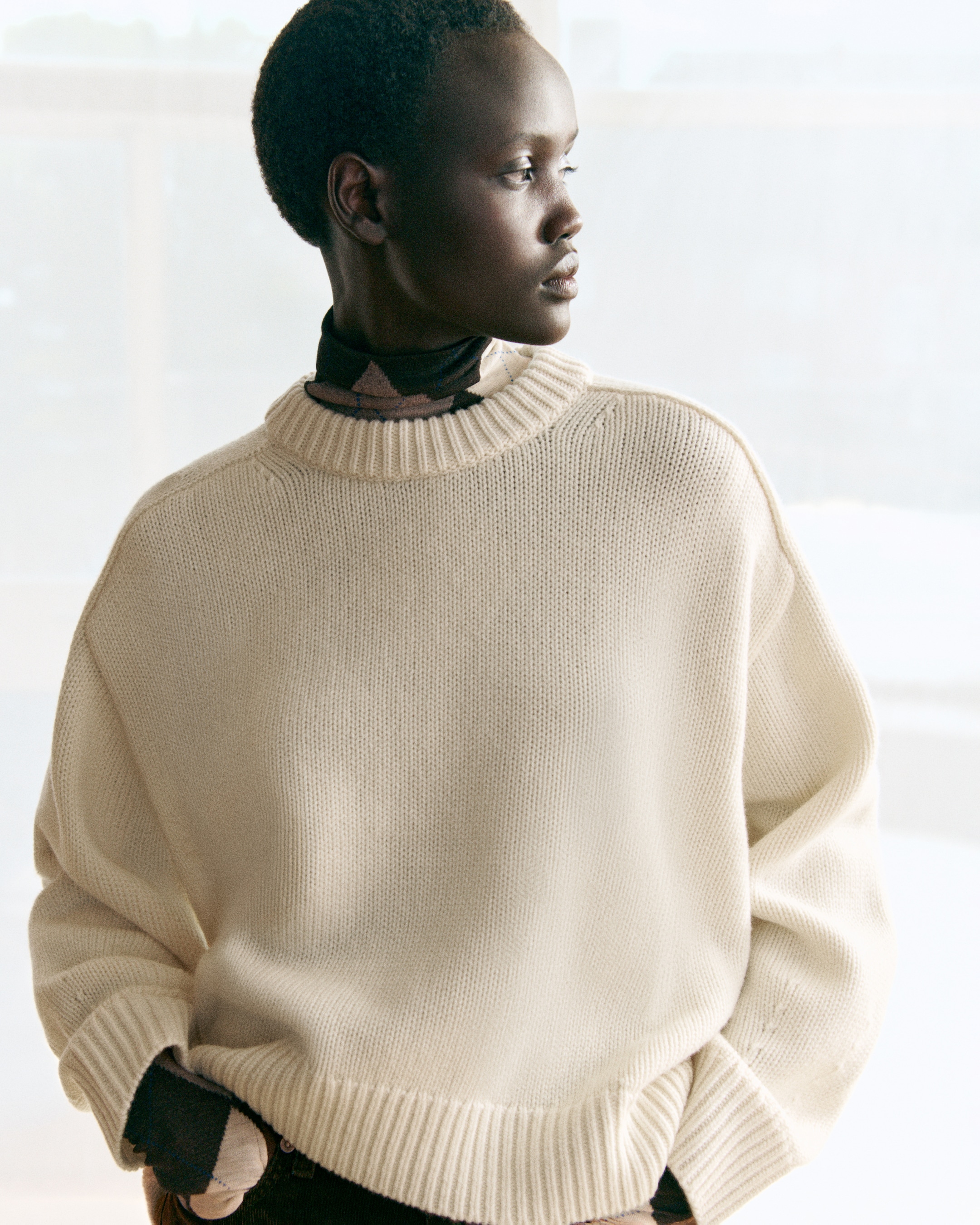 Person in a cream-colored sweater gazes to the side in a softly lit room, hands in pockets, looking thoughtful.