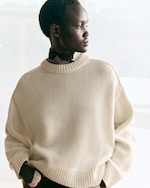Placeholder for: Person in a cream-colored sweater gazes to the side in a softly lit room, hands in pockets, looking thoughtful.
