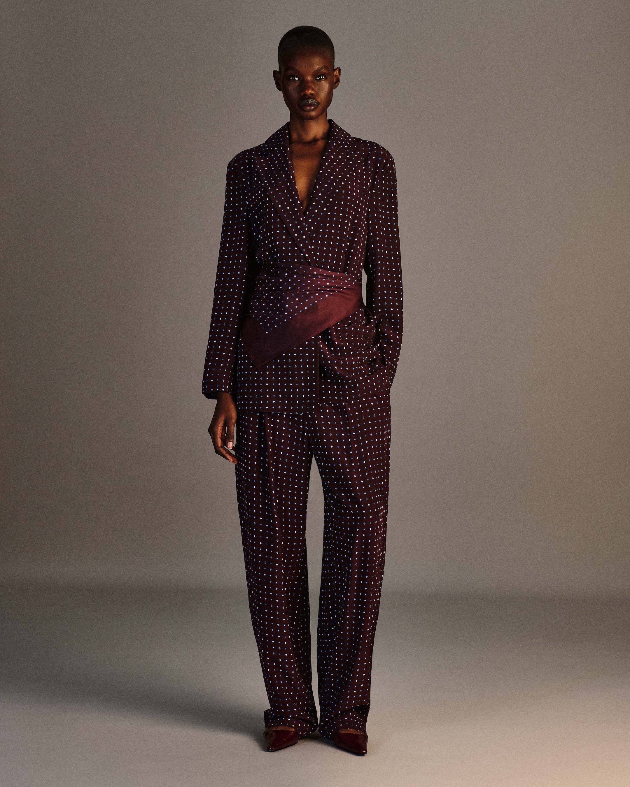 Person in a polka dot suit with a purple sash stands against a neutral background, projecting elegance and style.