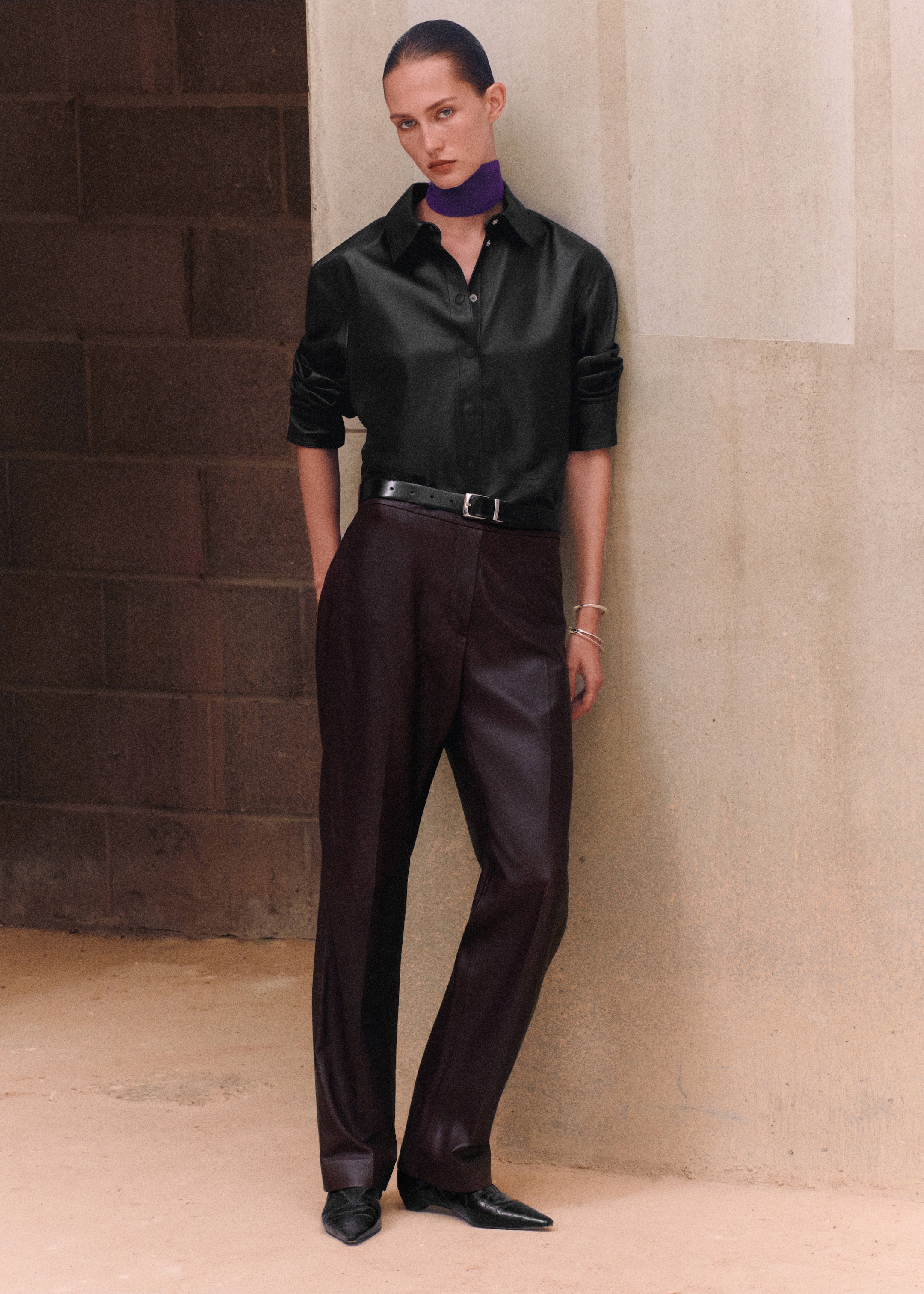 Person wearing a black shirt, dark trousers, and a purple scarf stands against a textured wall, with arms at sides and hands in pockets.