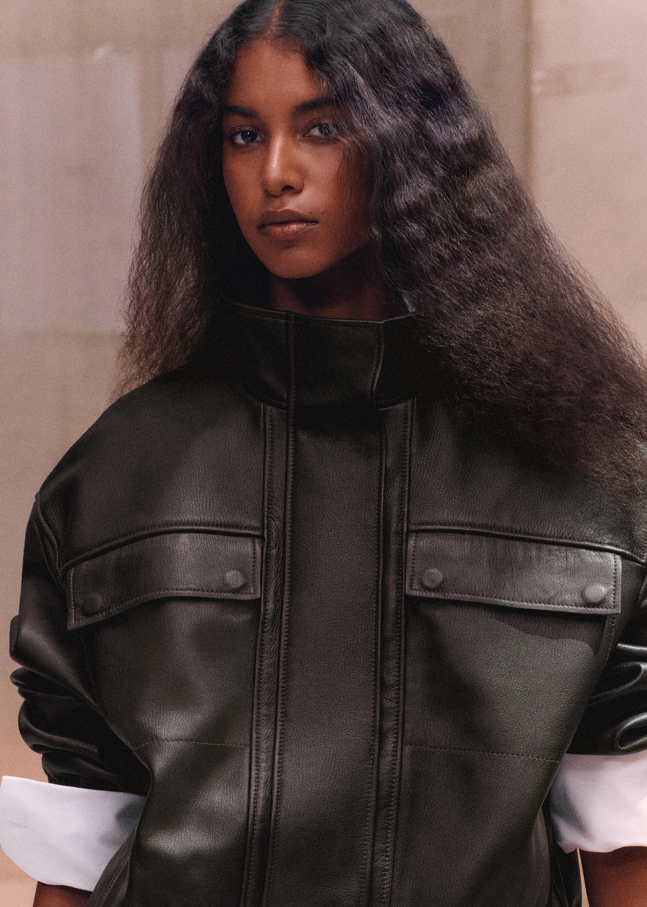 Person with long wavy hair wearing a high-collared, black leather jacket with buttoned pockets, against a neutral background.