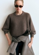Placeholder for: Person wearing a brown sweater and sunglasses, with a gray sweater tied around their waist, standing against a plain background.