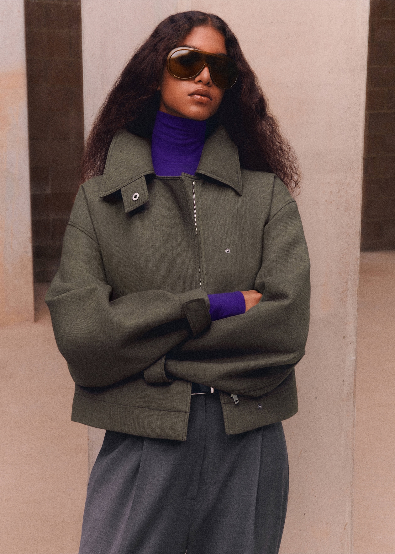 A person with long curly hair wearing a green jacket, purple turtleneck, and large sunglasses stands confidently with arms crossed.
