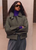 Placeholder for: A person with long curly hair wearing a green jacket, purple turtleneck, and large sunglasses stands confidently with arms crossed.