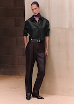 Placeholder for: Person leaning against a wall in a black shirt, dark trousers, and black shoes, with a purple scarf around the neck. Neutral expression.