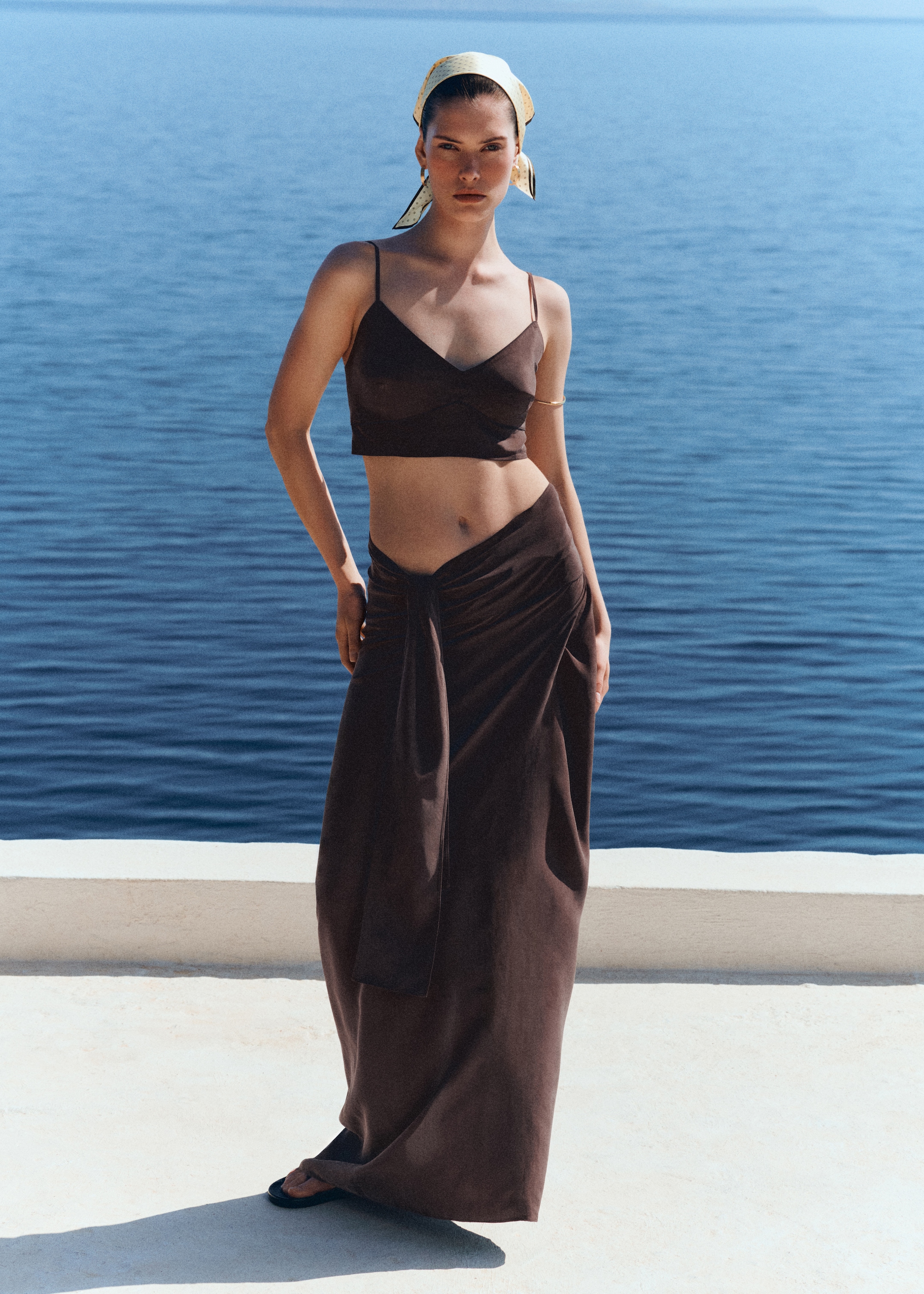 Woman by the sea wearing a dark brown crop top and long skirt, with a headscarf and sandals, against a blue ocean backdrop.