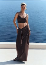 Placeholder for: Woman by the sea wearing a dark brown crop top and long skirt, with a headscarf and sandals, against a blue ocean backdrop.