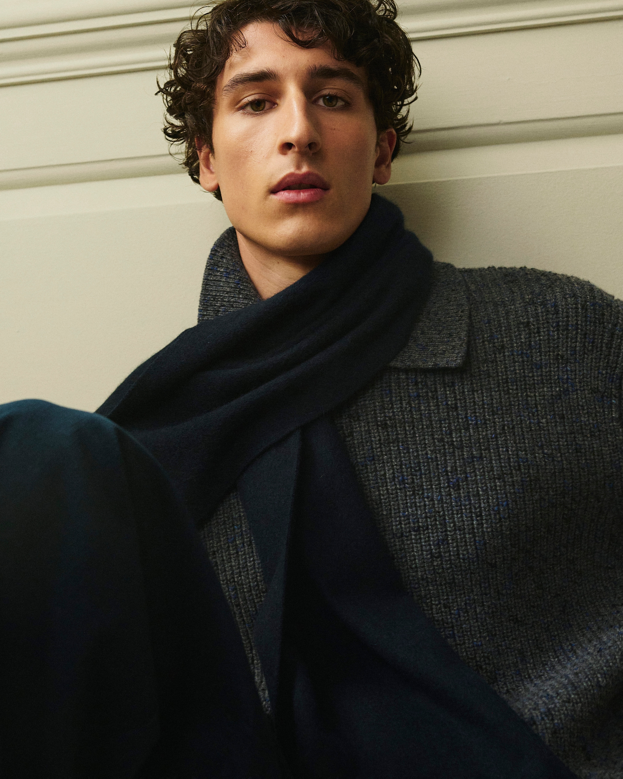 A person with curly hair wearing a dark scarf and a wool sweater, seated against a neutral background.