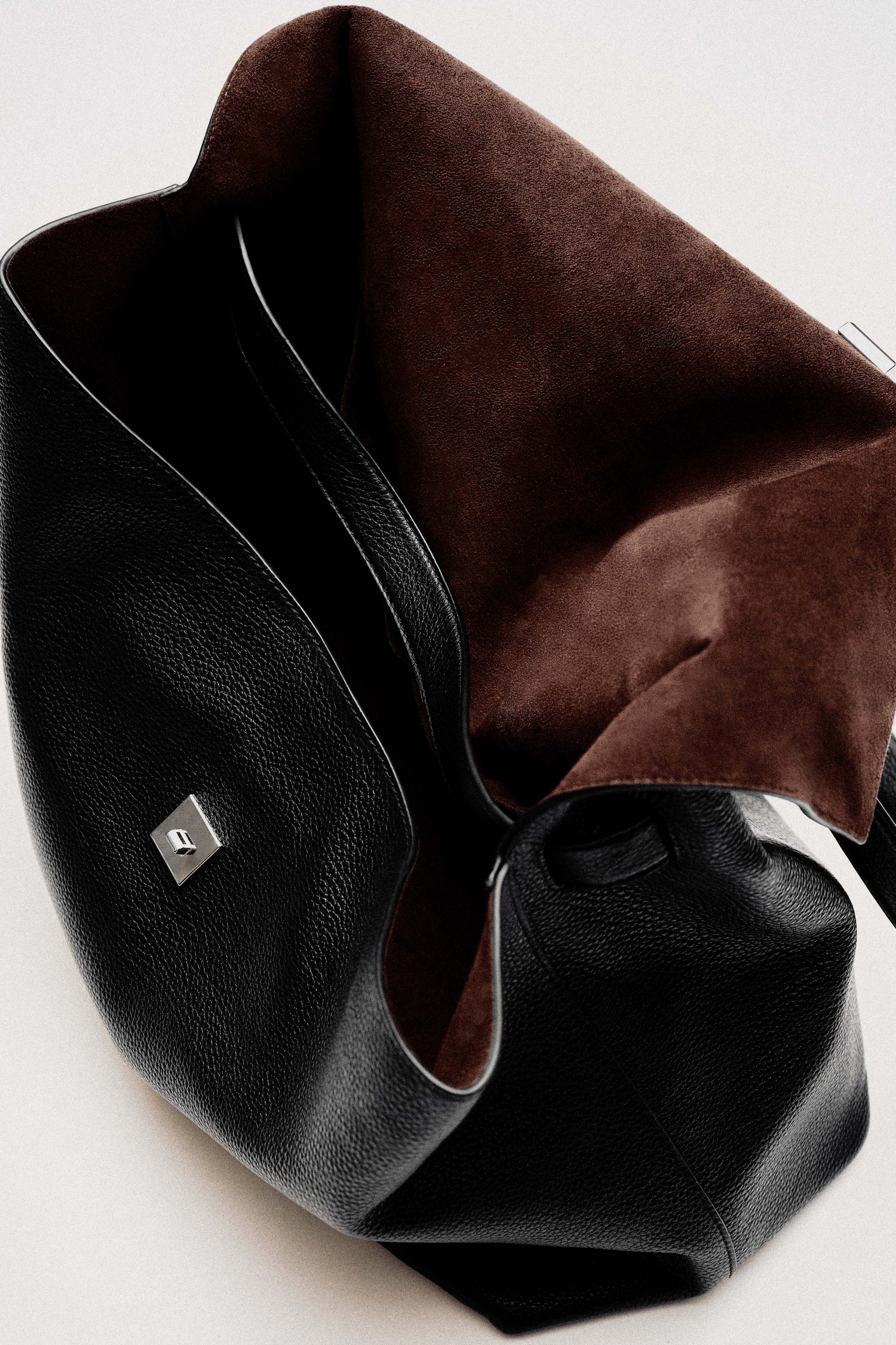 Black leather handbag with an open top revealing a brown suede interior, featuring a small silver square clasp on the exterior.