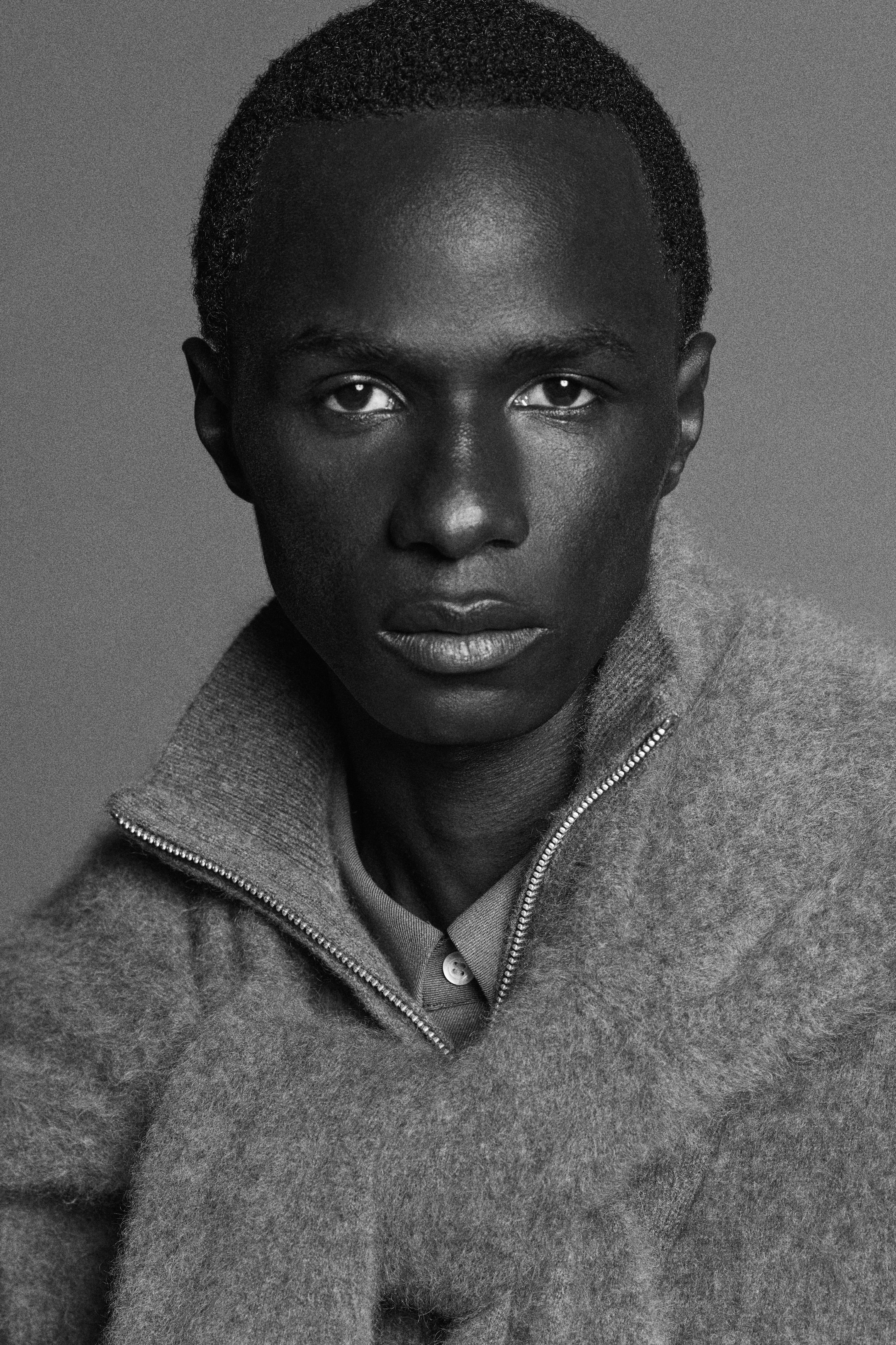 Black and white portrait of a person with short hair, wearing a textured sweater with a zipper, looking directly at the camera.