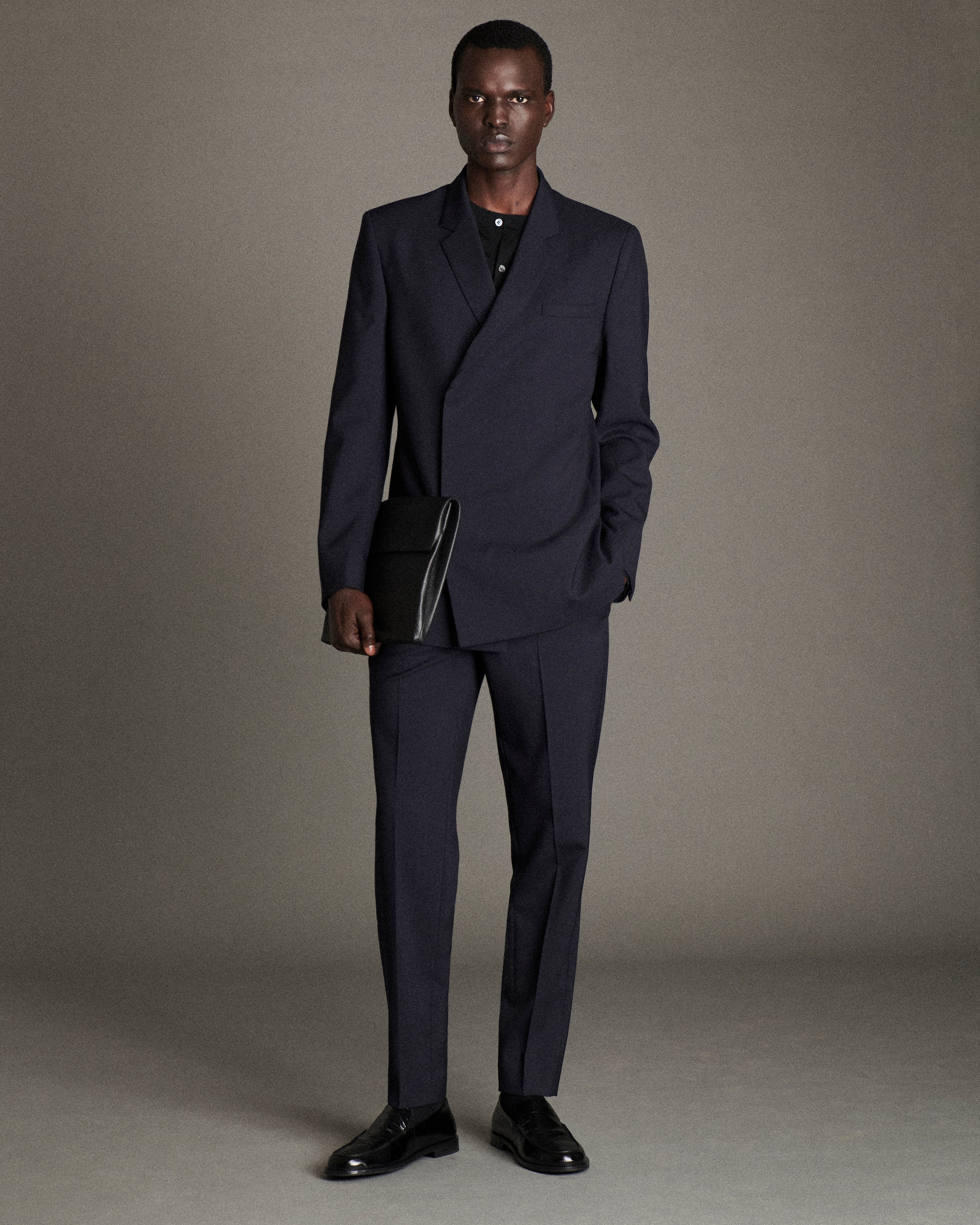 A person in a dark, tailored suit stands holding a large black folder, with one hand in their pocket, on a neutral backdrop.