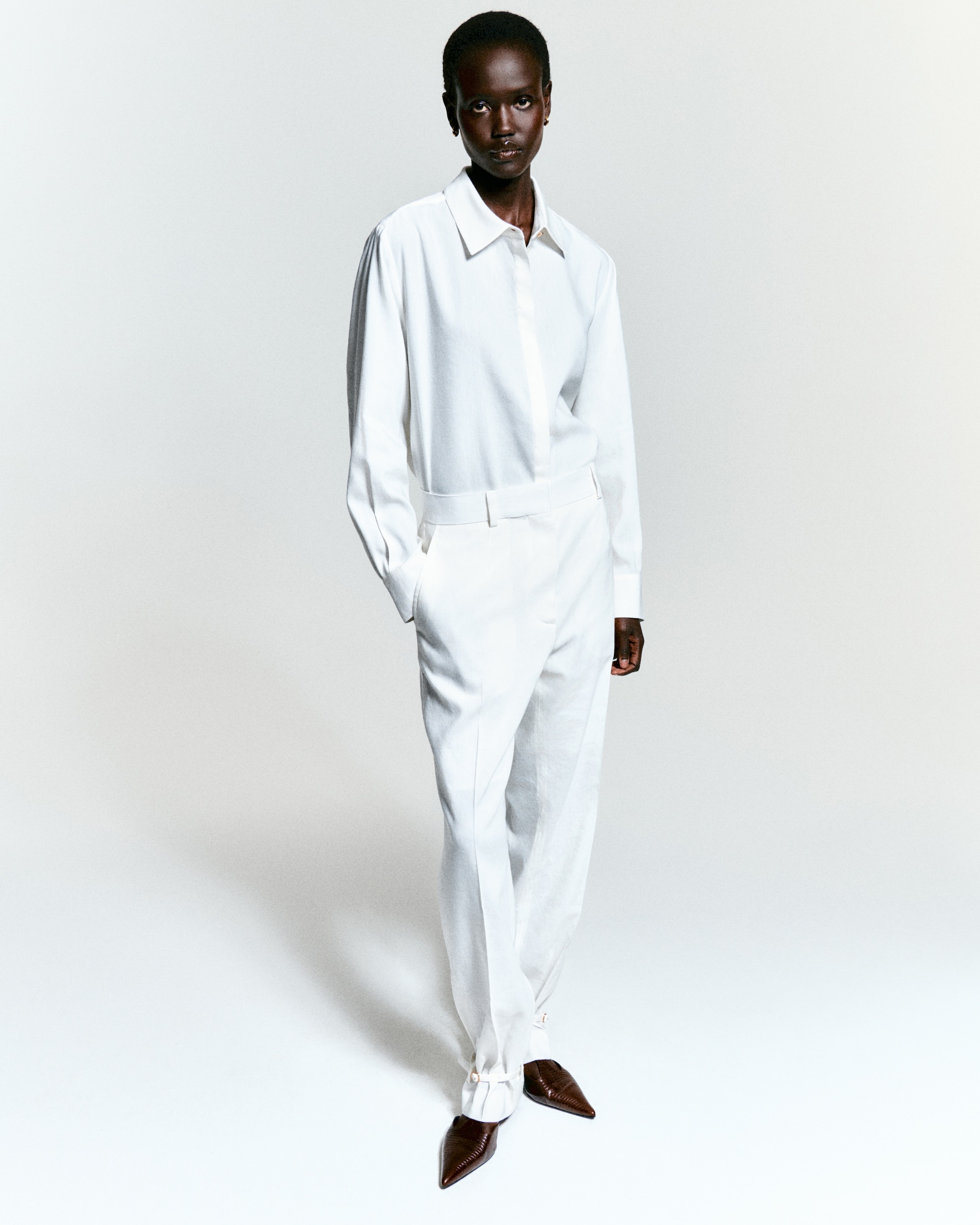 Person in a stylish white outfit with a collared shirt and high-waisted pants, paired with pointed brown shoes, standing confidently.