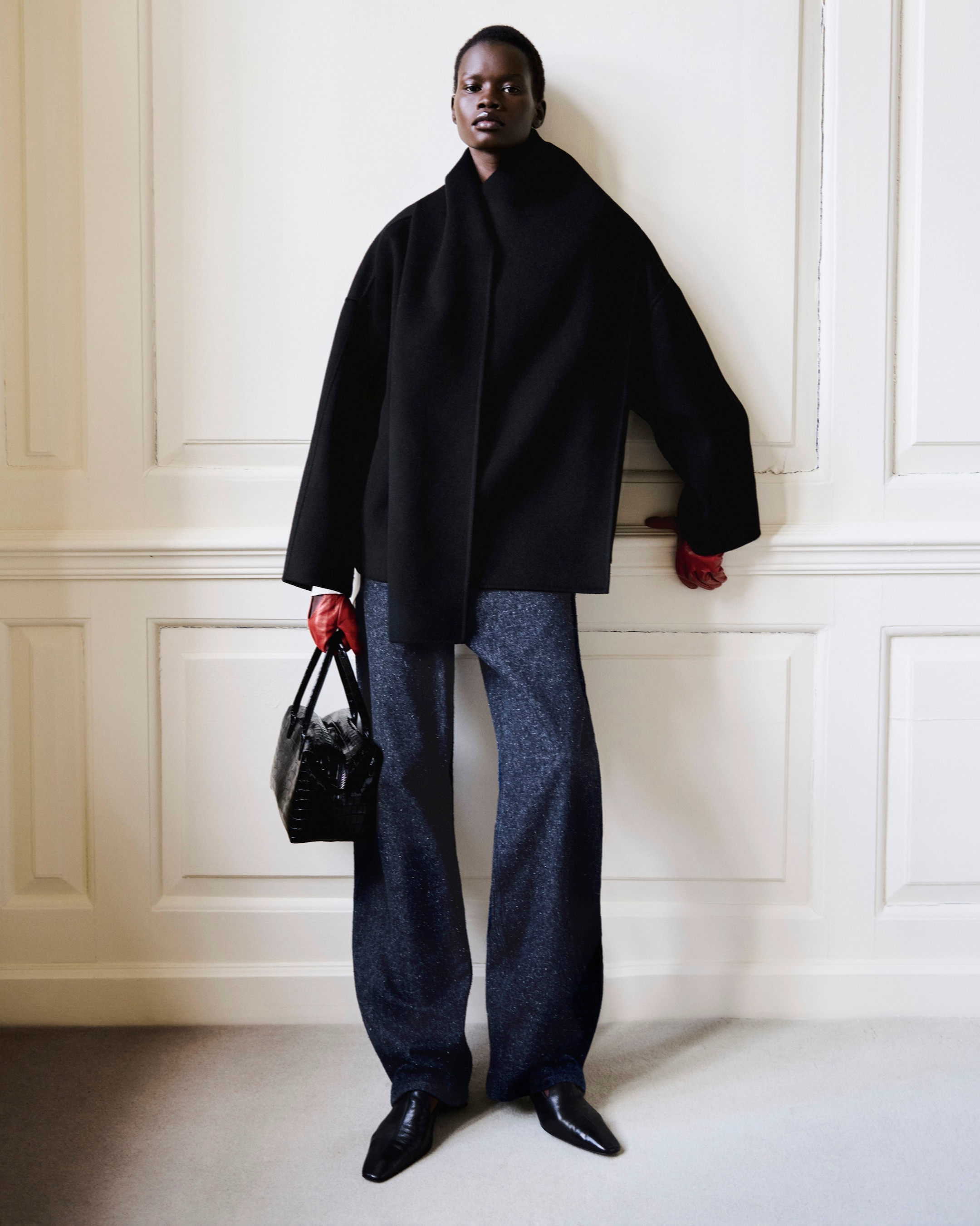 Person wearing a black coat, wide-legged blue trousers, holding a black handbag with red gloves, standing indoors by a white wall.