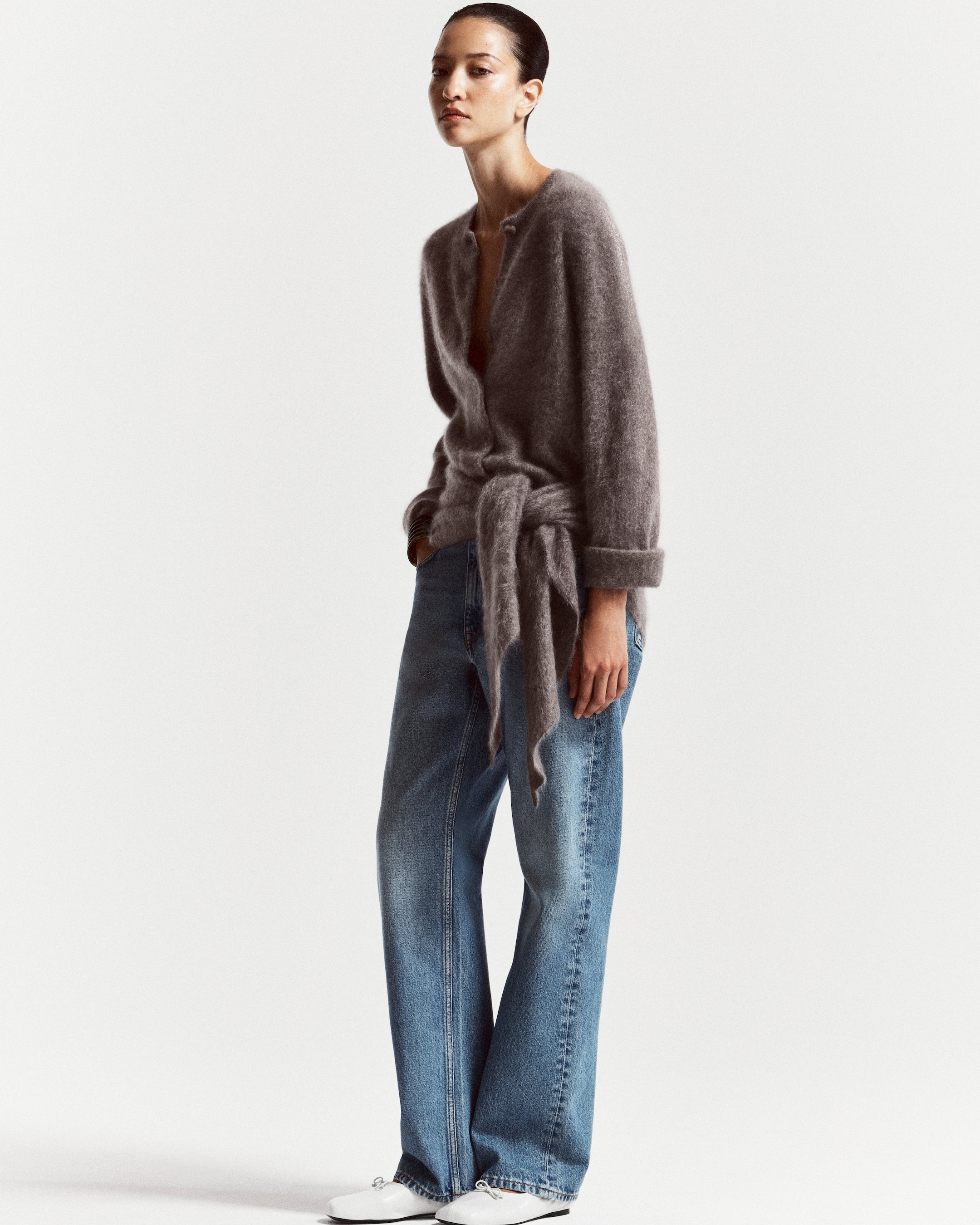 A person wearing a gray fuzzy cardigan with a belt, blue wide-leg jeans, and white shoes, posing against a plain white background.