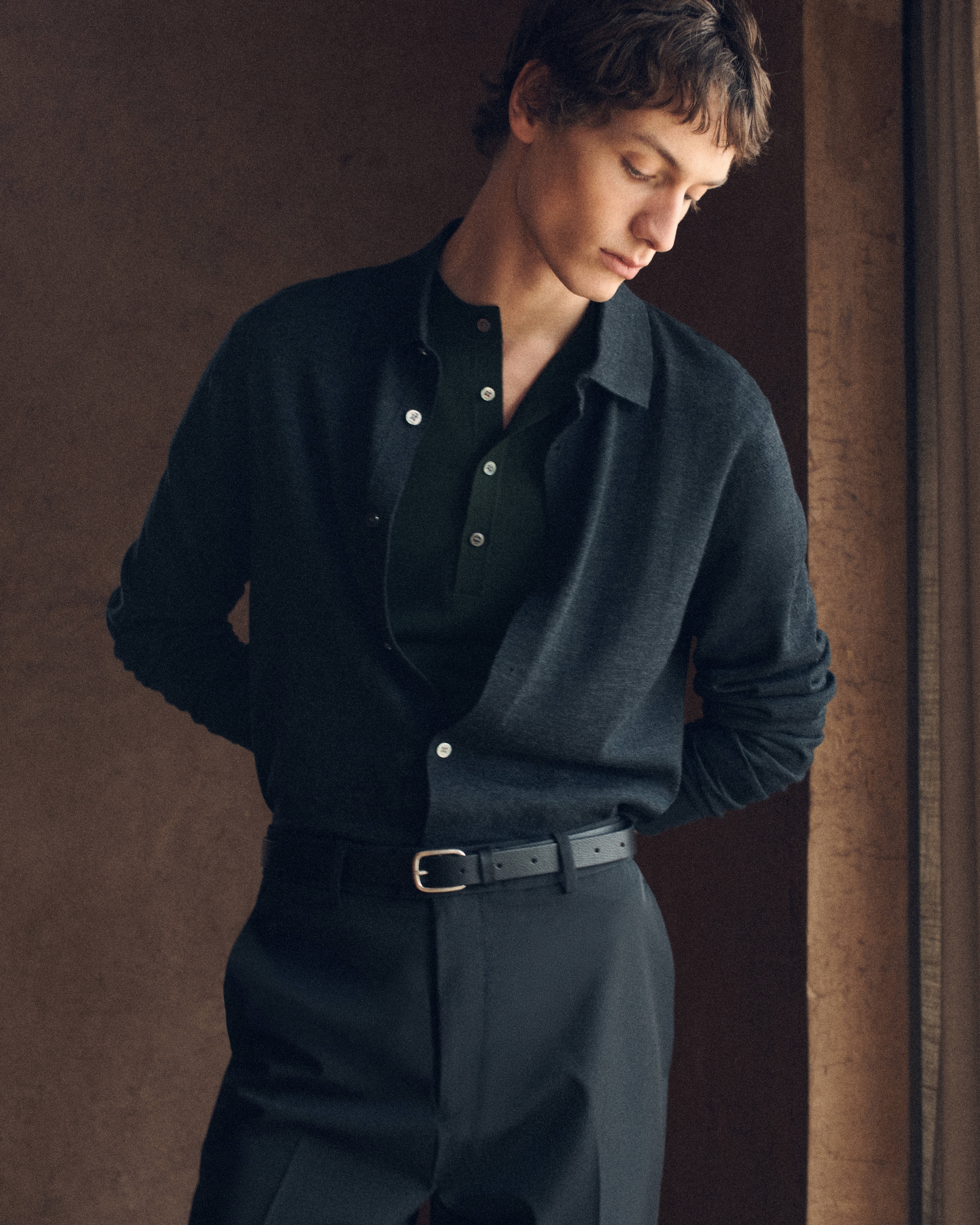 A person in a dark shirt and trousers stands indoors, looking down with hands behind their back, in soft lighting.