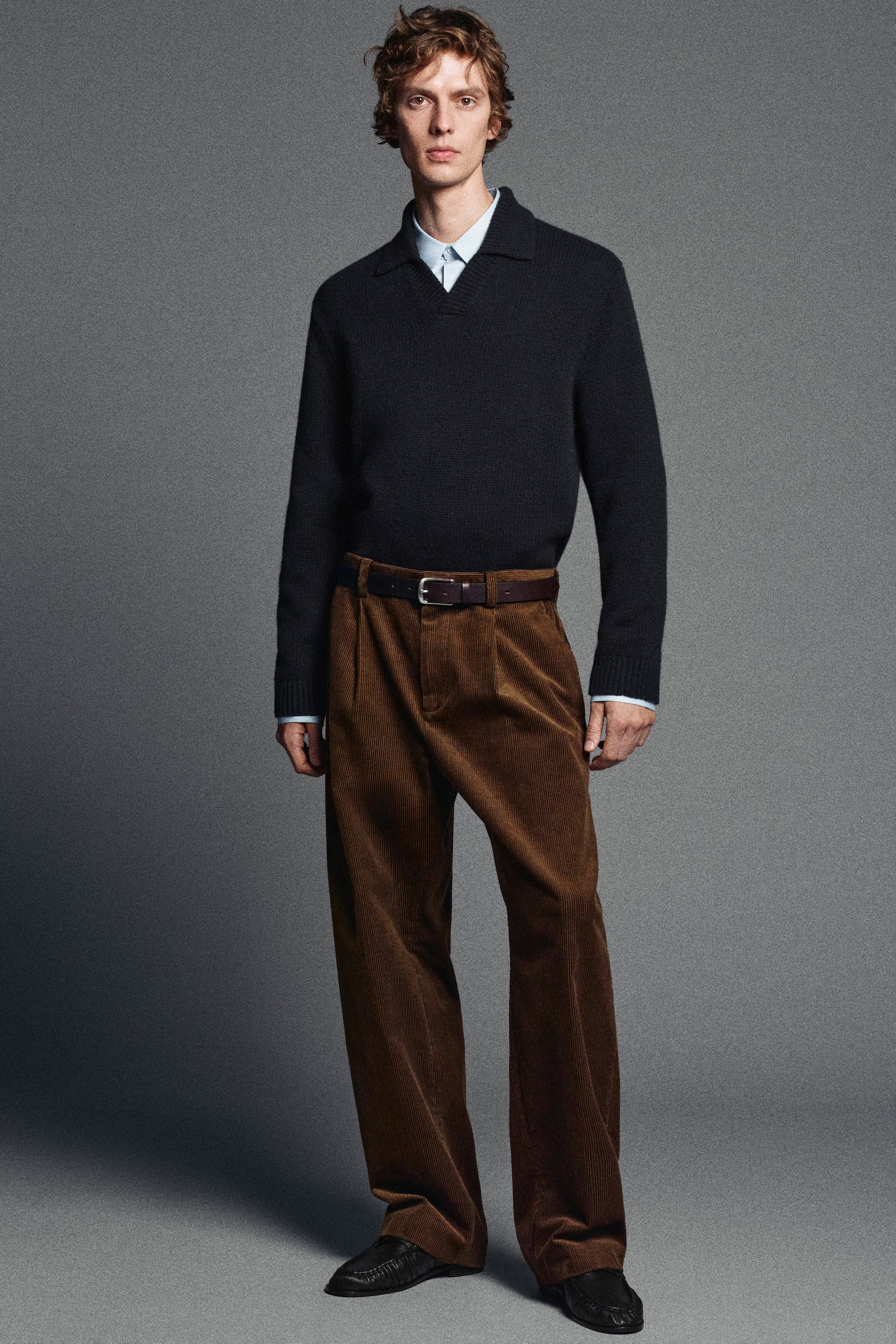 A person wearing a dark sweater over a light shirt, with brown corduroy pants and black shoes, stands against a gray background.