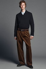 Placeholder for: A person wearing a dark sweater over a light shirt, with brown corduroy pants and black shoes, stands against a gray background.
