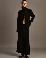 Placeholder for: A person in a black buttoned outfit with a high collar and long skirt, posing with one hand in their pocket against a neutral background.