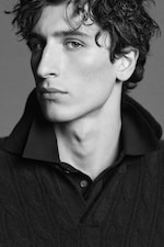 Placeholder for: Black and white portrait of a person with curly hair, wearing a dark collared shirt and sweater, gazing into the distance.