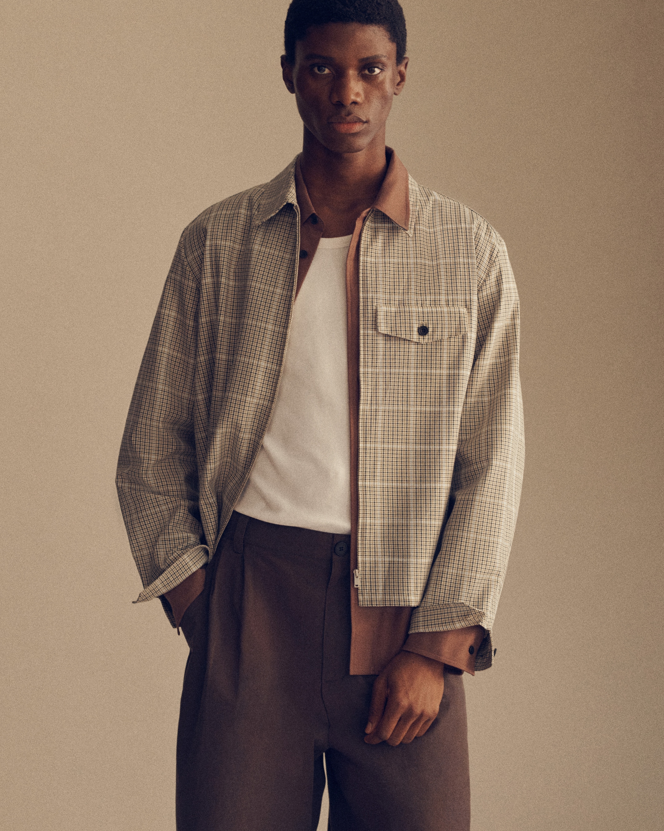 A man wearing a plaid jacket over a white shirt and brown pants, standing against a plain background.