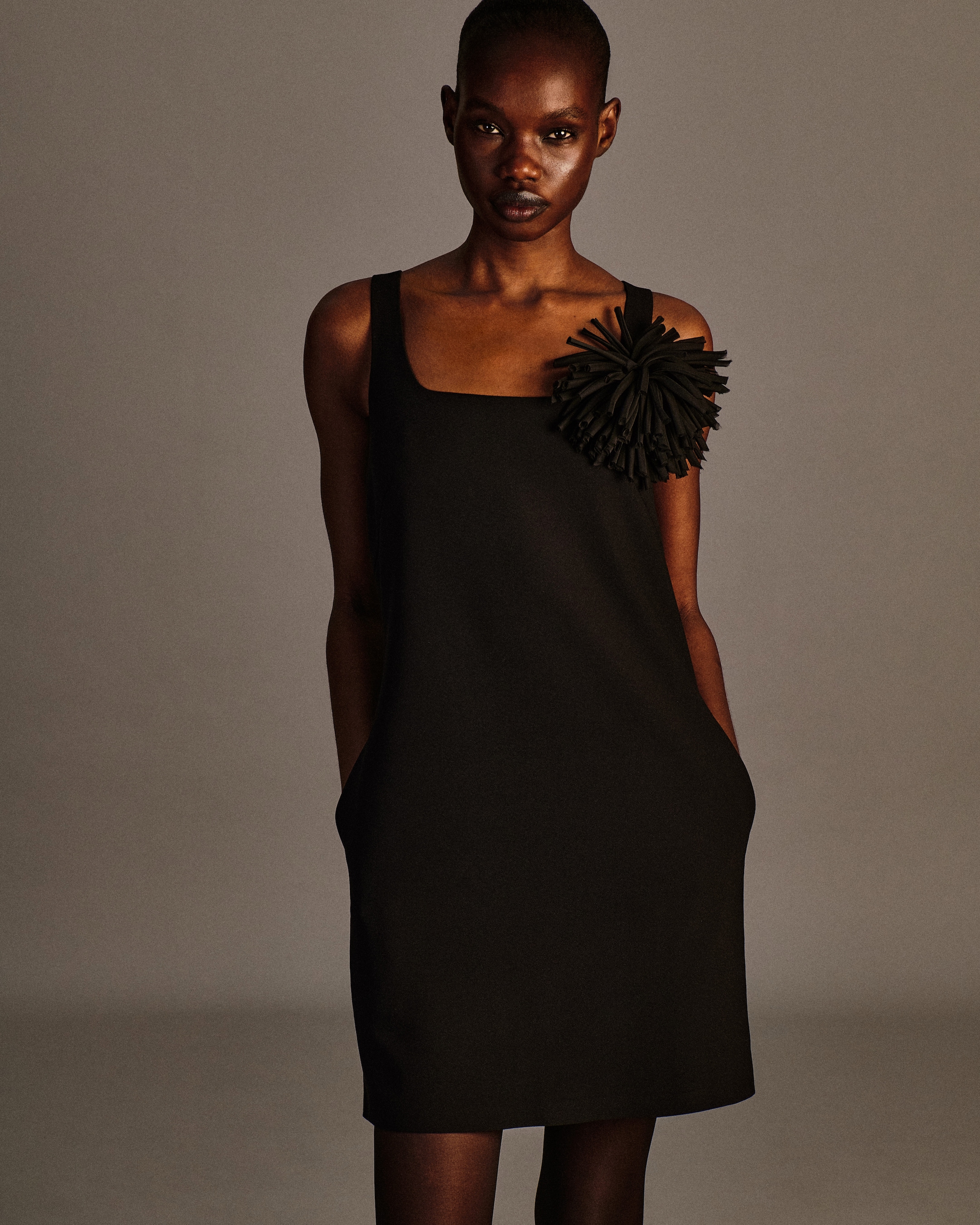 A person wearing a black dress with a large decorative flower on the shoulder stands against a neutral background.