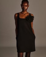 Placeholder for: A person wearing a black dress with a large decorative flower on the shoulder stands against a neutral background.