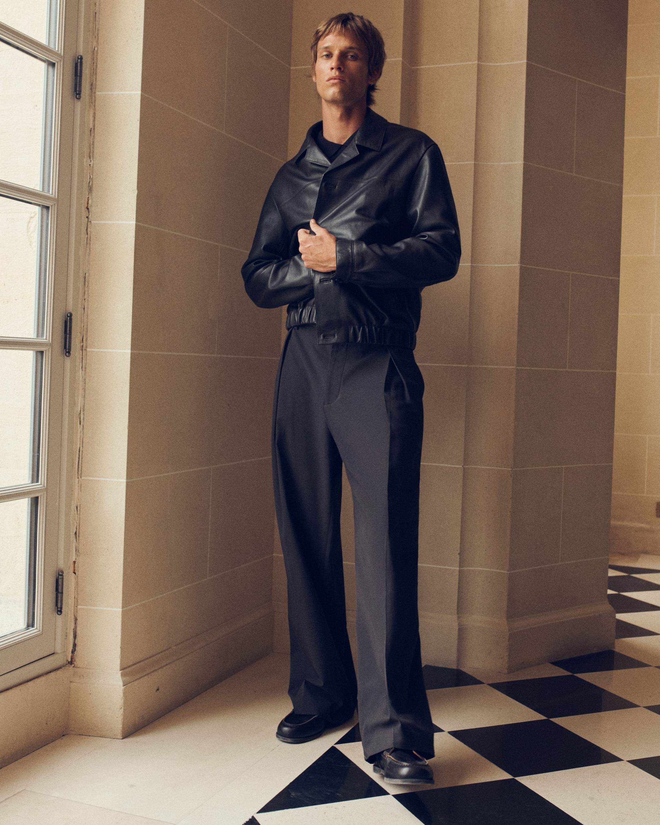 A person in a black leather jacket and wide black trousers stands in a sunlit room with checkered floor tiles and beige walls.