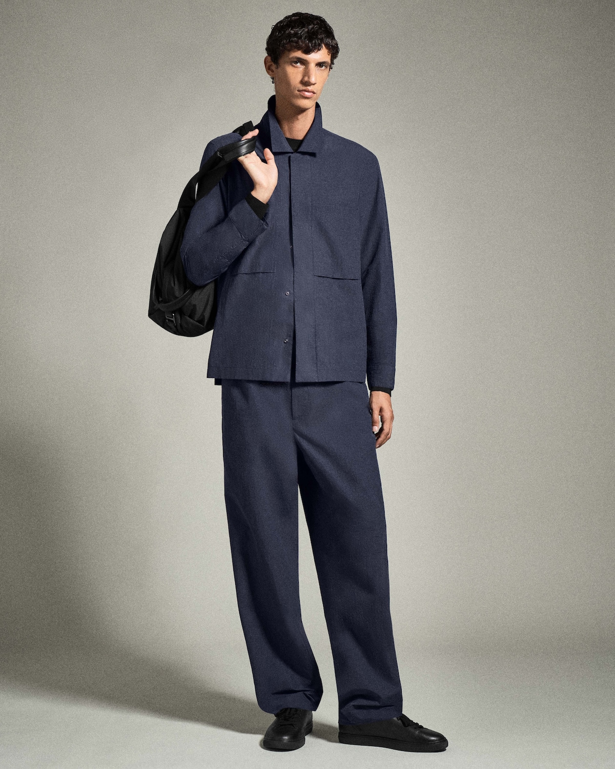 Man in a dark blue suit holds a black bag over his shoulder, standing against a neutral background.