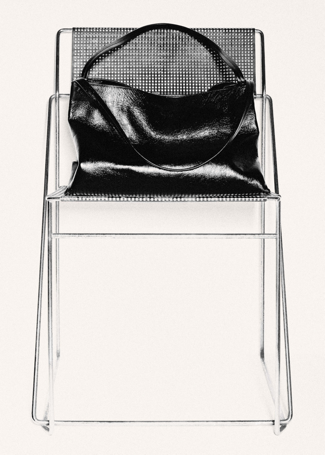 A shiny black handbag placed on a modern wireframe chair with a perforated metal seat and backrest.