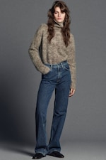 Placeholder for: A person wearing a fuzzy gray turtleneck sweater and blue jeans stands with one hand in their pocket against a gray background.