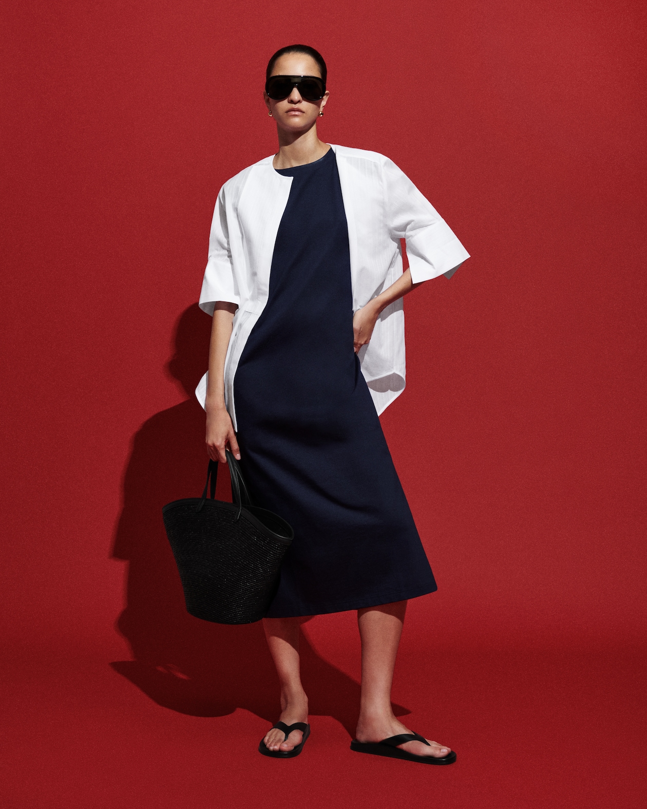 Red background with mauve dress with white button up and sandals with large oversized sunglasses