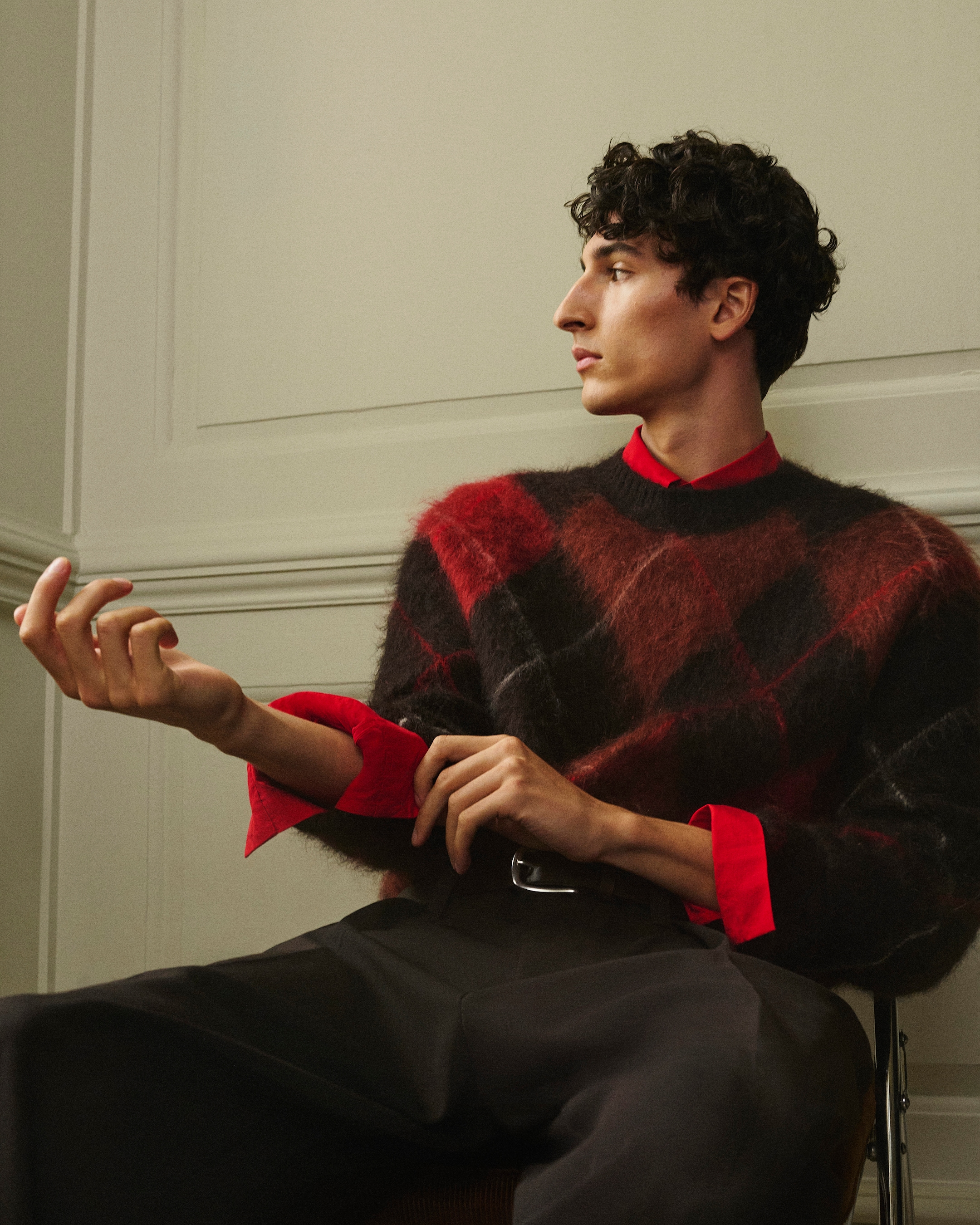 Person seated, wearing a red and black argyle sweater over a red shirt, looking to the side, with curly dark hair and expressive hand gesture.
