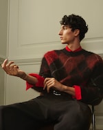 Placeholder for: Person seated, wearing a red and black argyle sweater over a red shirt, looking to the side, with curly dark hair and expressive hand gesture.