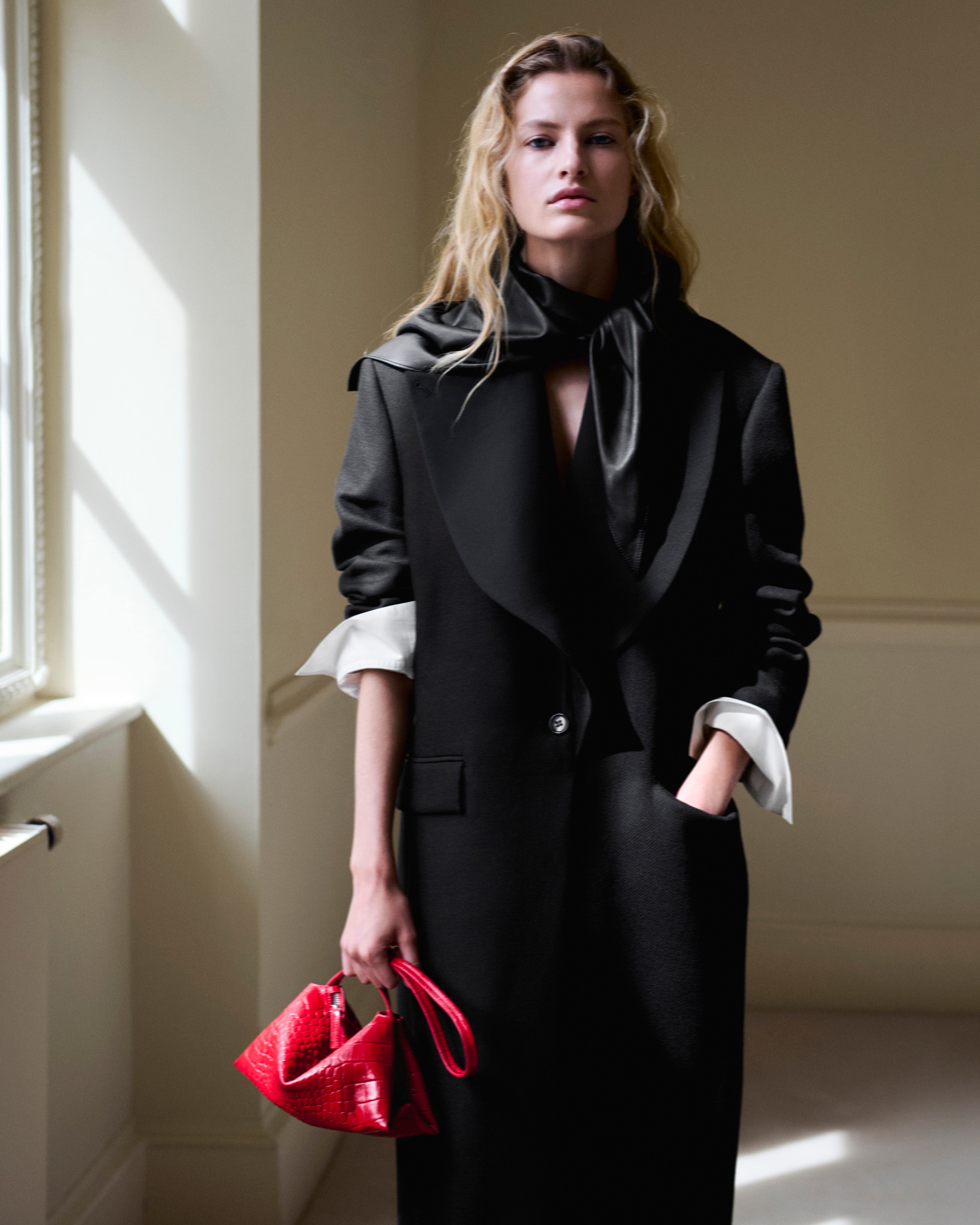 A person with long blond hair wearing a black coat and scarf, holding a small red handbag, standing in a well-lit room.