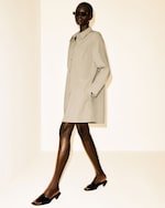 Placeholder for: Person wearing a beige long coat, sunglasses, and black heeled sandals, walking in a minimalistic bright space.