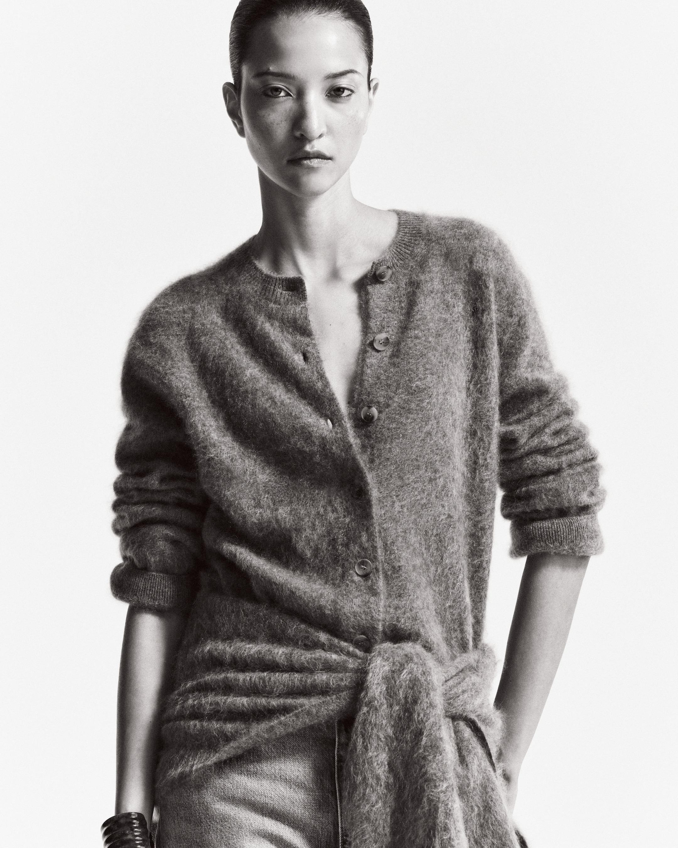 A monochrome image of a person wearing a fuzzy buttoned cardigan and jeans, with hair tied back, gazing directly at the camera.