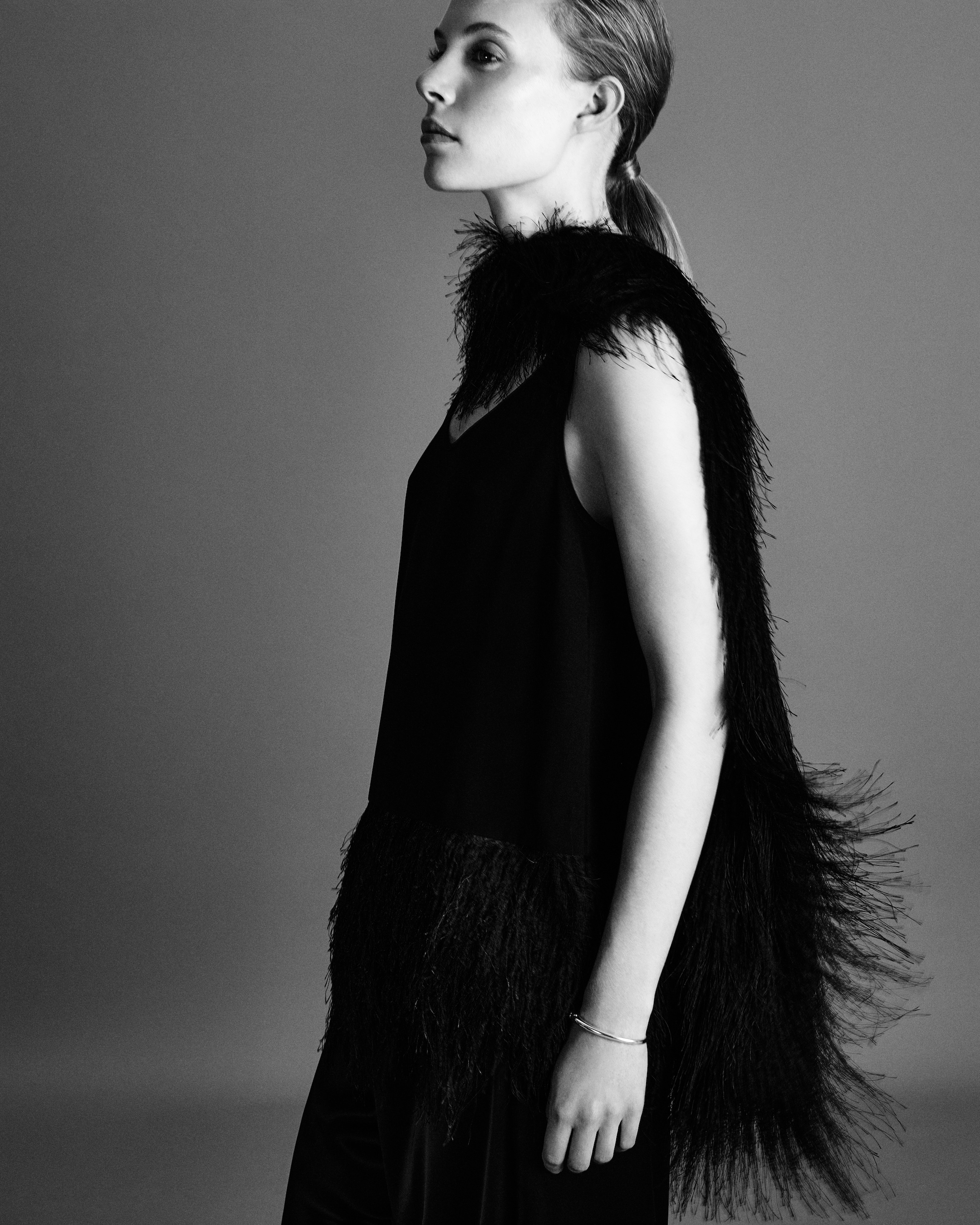 A monochrome side profile of a person wearing a black outfit with feathered details, their hair pulled back, against a gray background.