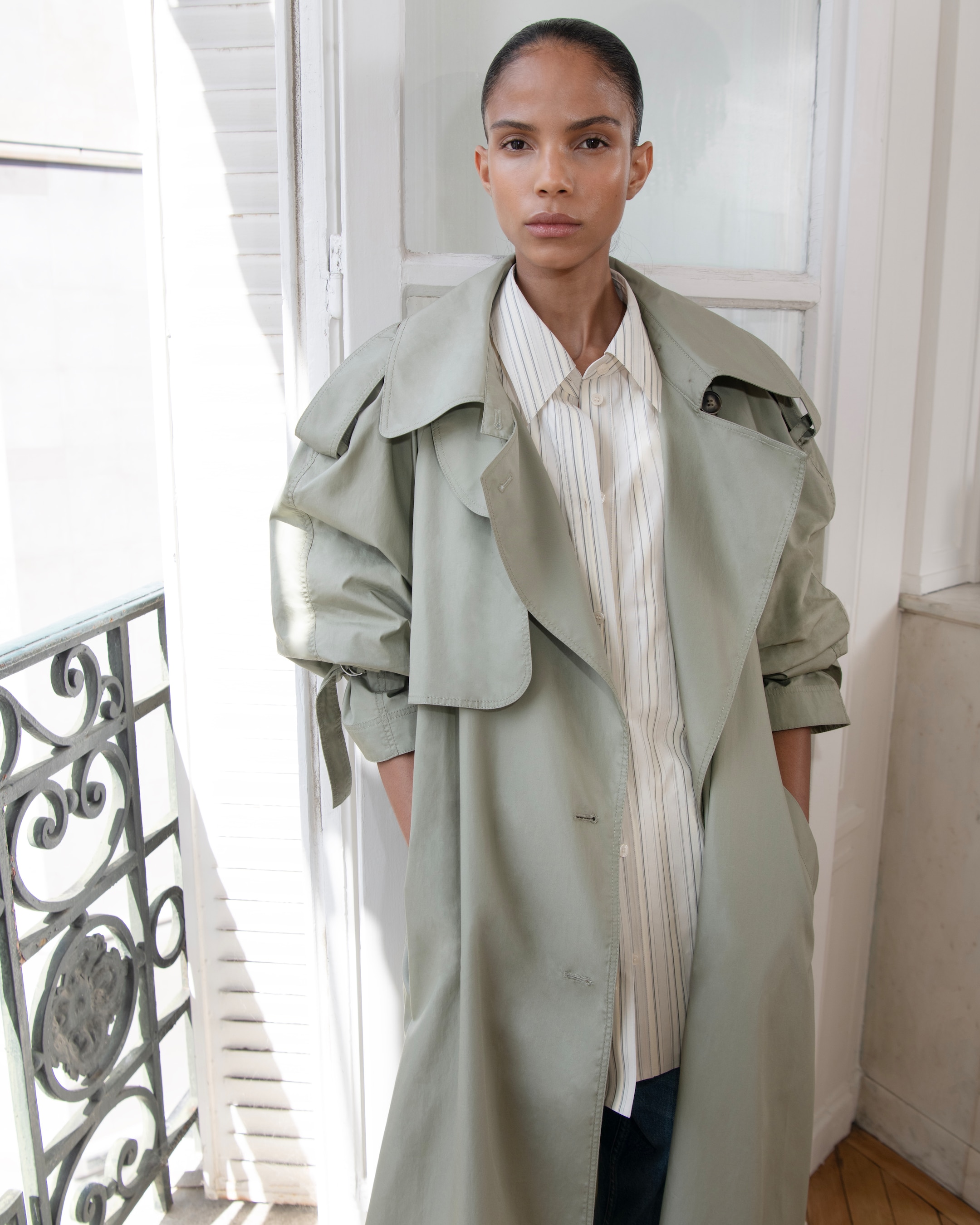 A person with a neutral expression stands indoors wearing a light green trench coat over a striped shirt, with their hands in their coat pockets.
