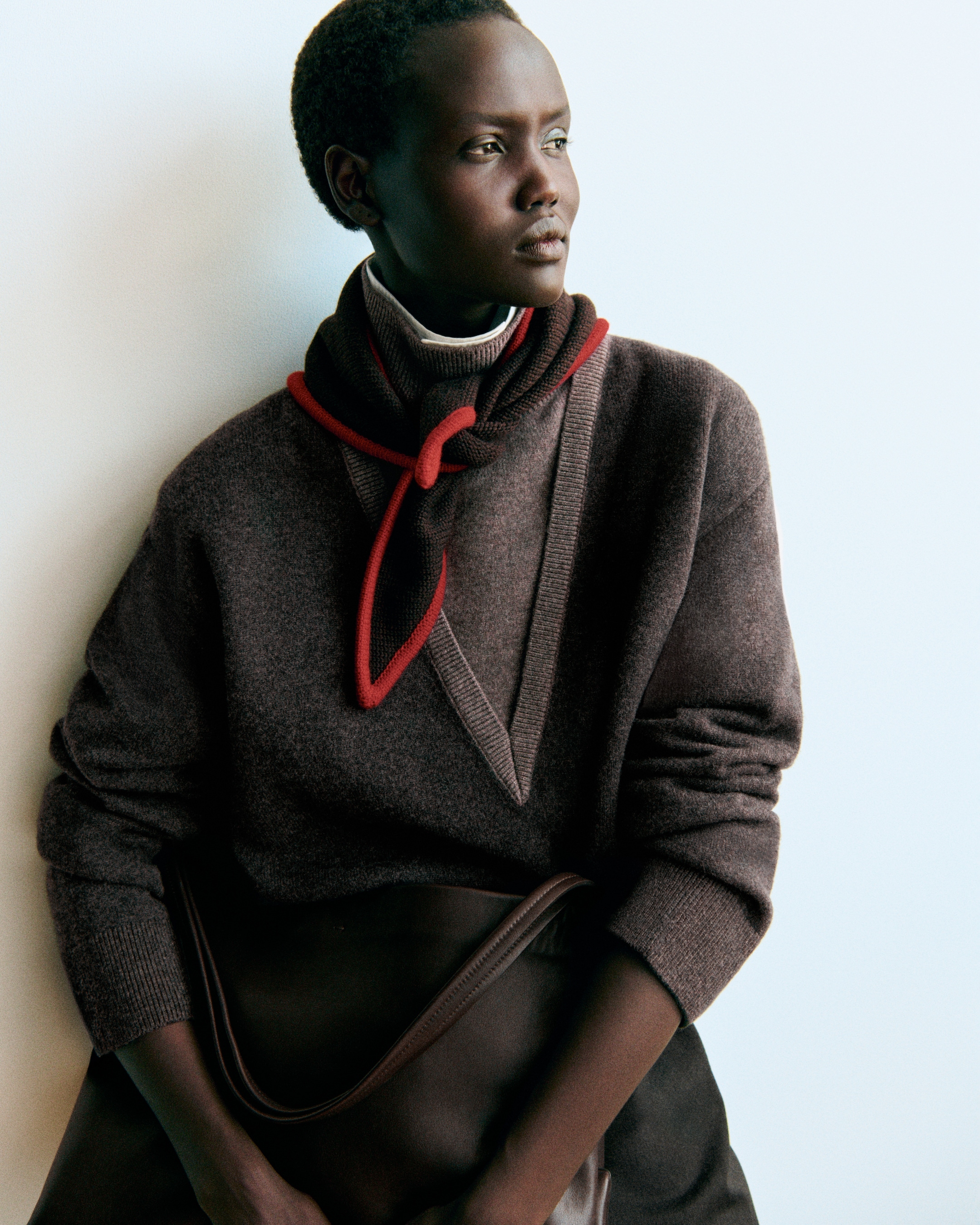 A person in a dark brown sweater with a red-trimmed scarf, looking thoughtfully to the side against a light background.