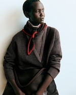 Placeholder for: A person in a dark brown sweater with a red-trimmed scarf, looking thoughtfully to the side against a light background.