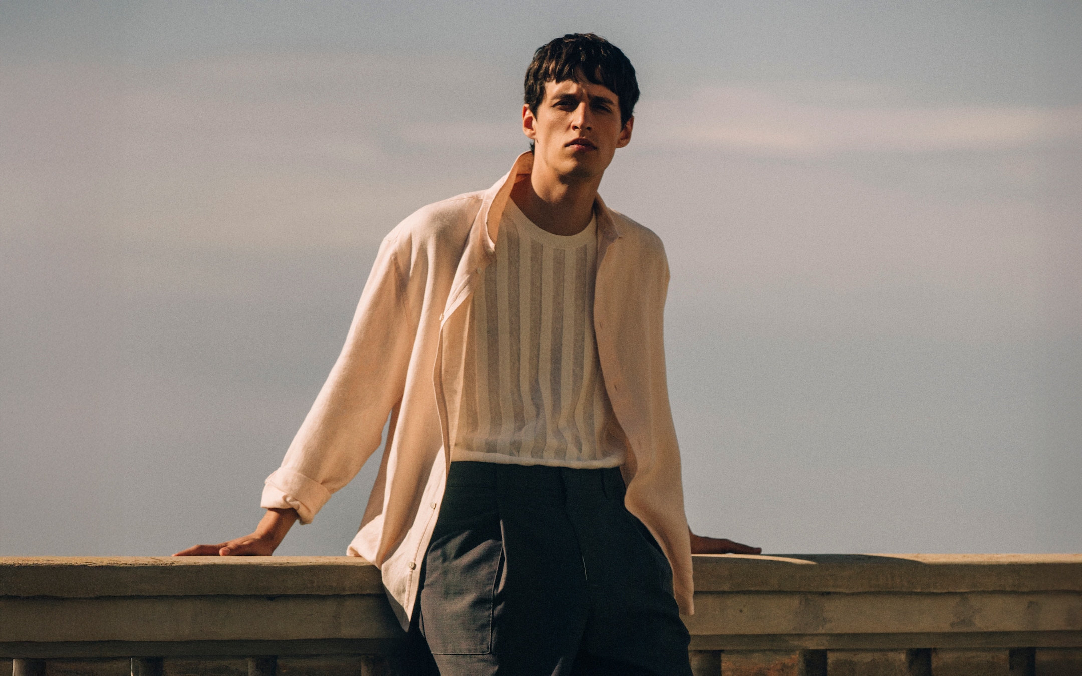 A sunlit, medium shot of a man standing outdoors and leaning against a stone railing. He wears a light beige, long-sleeved button-down shirt left open over a white, vertical-striped knit T-shirt tucked into dark gray tailored trousers. The background features a soft, hazy sky, emphasizing a relaxed and sophisticated summer holiday aesthetic.