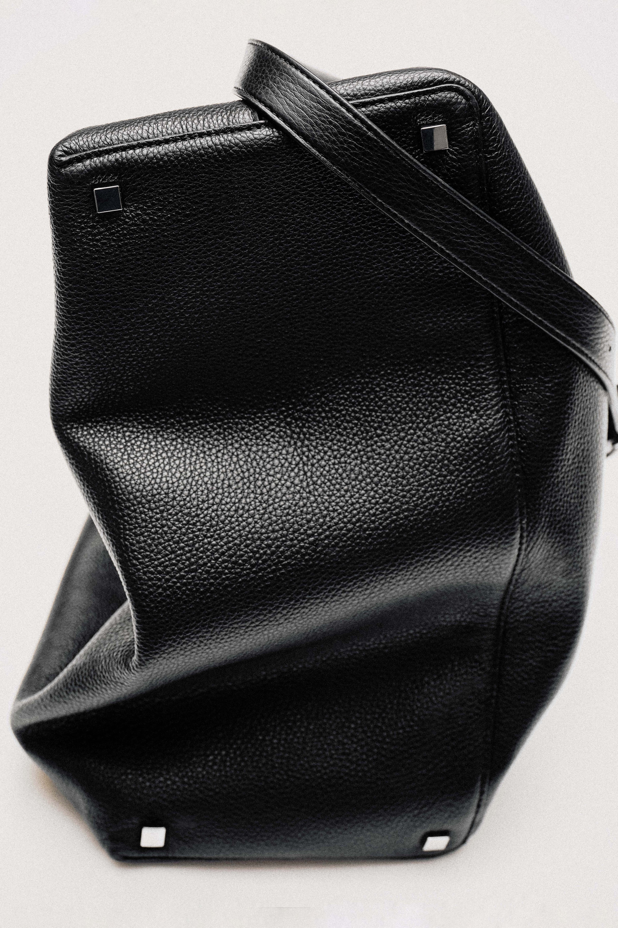Black textured leather handbag with a single strap, square metal accents on the base, placed on a light background.