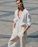 Placeholder for: Person wearing a white open-neck shirt and beige trousers stands outdoors on a sunny day with hands in pockets.